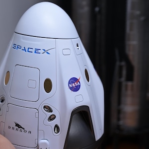 May include: A white model of a SpaceX Dragon spacecraft with the words "SpaceX" and "Dragon" printed on the side. The spacecraft has a black nose cone and a NASA logo on the side.