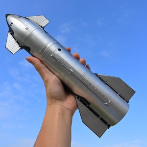 May include: A silver and black model of a spacecraft with a pointed nose and a set of wings. The model is held in a person's hand against a blue sky.