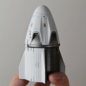 May include: A white and black model of a spacecraft capsule. The capsule has a rounded top with detailed panel lines, small windows, and gold-colored accents. The lower section is black and white with a textured surface.