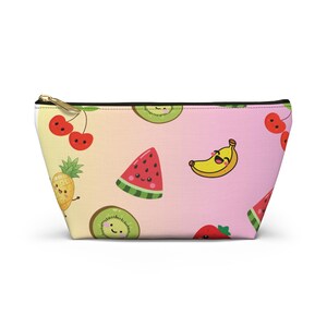 Può includere: Una pochette con cerniera caratterizzata da un motivo ripetuto di frutta cartoon sorridente, tra cui ciliegie, un ananas, una fetta di anguria, una banana, un kiwi e una fragola su uno sfondo sfumato dal giallo al rosa con cursore dorato.