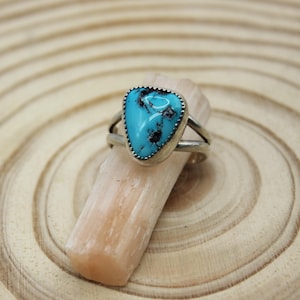 May include: A silver ring with a triangular, turquoise stone. The stone has dark blue and black inclusions and is set in a scalloped bezel. The ring sits on a light-colored, rectangular object against a wood grain background.