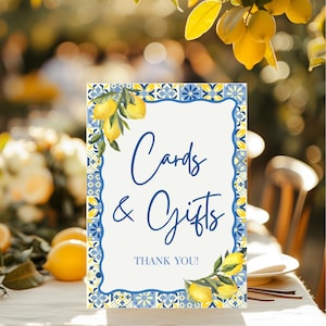 May include: A rectangular sign with a white background and blue border, featuring the words "Cards & Gifts" in elegant script. The sign is decorated with lemon illustrations and the words "THANK YOU!" at the bottom. The sign is placed on a table.