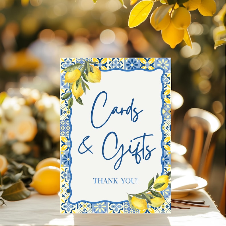 May include: A rectangular sign with a white background and blue border, featuring the words "Cards & Gifts" in elegant script. The sign is decorated with lemon illustrations and the words "THANK YOU!" at the bottom. The sign is placed on a table.