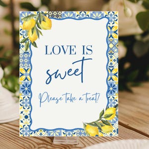 May include: A rectangular sign with a white background and a blue and yellow decorative border. The sign reads "LOVE IS sweet Please Take a Treat!" and features illustrations of lemons.