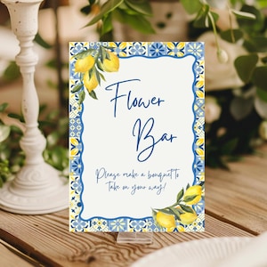 May include: A decorative sign with a white background and blue border, featuring the text "Flower Bar." The sign is adorned with lemon illustrations and the phrase "Please make a bouquet to take on your way!" It is displayed on a wooden surface.