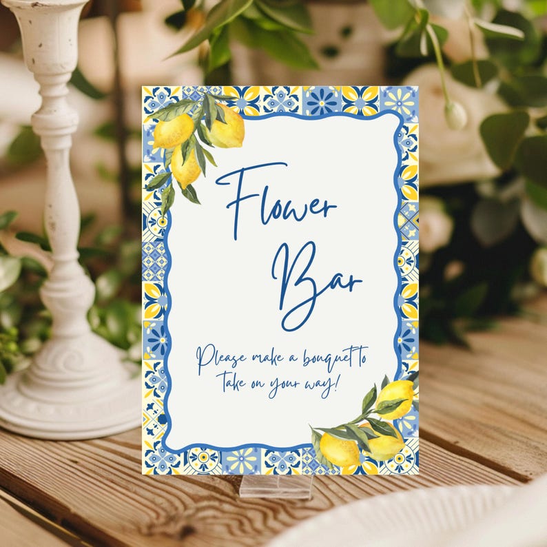 May include: A decorative sign with a white background and blue border, featuring the text "Flower Bar." The sign is adorned with lemon illustrations and the phrase "Please make a bouquet to take on your way!" It is displayed on a wooden surface.