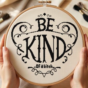 May include: A circular wooden embroidery hoop containing a piece of cream-colored Aida fabric. The fabric features black cross-stitched text that reads "BE KIND" in large, stylized letters, with "OF A BITCH" in smaller text below, framed by decorative swirls.
