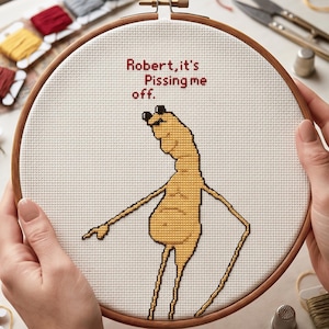 Pode incluir: Um bastidor de bordado em ponto cruz com o texto "Robert, it's Pissing me off." O desenho apresenta uma figura de desenho animado amarela com braços longos e um dedo apontando. Fios de bordar e tesouras são visíveis.