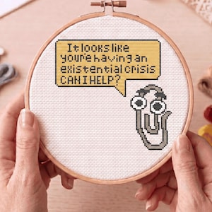 May include: A cross-stitch embroidery hoop with a humorous design. The design features a speech bubble with the text "It looks like you're having an existential crisis. CAN I HELP?" and a pixelated paperclip character.