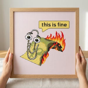 May include: A framed cross-stitch embroidery featuring a silver paperclip character with large eyes standing on a yellow document that is engulfed in orange and red flames, with a yellow speech bubble containing the black text this is fine.