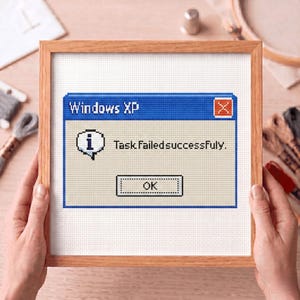 Op de afbeelding: Kruissteekkunst in een houten lijst. Het kunstwerk toont een Windows XP-foutmeldingsvenster met de tekst "Task Failed Successfully" en een "OK"-knop. De achtergrond is lichtbeige.