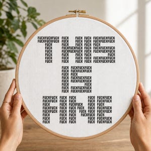 May include: A circular wooden embroidery hoop containing white fabric with black cross-stitched text. The text is arranged to spell out the phrase THIS IS FINE, with each letter composed entirely of the repeated word FUCK in a blocky font style.
