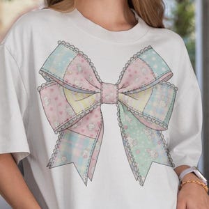 Puede incluir: Camiseta blanca con un gran lazo en colores pastel. El diseño del lazo incorpora secciones rosas, azules y amarillas con estampados florales y de corazones, delineadas con un ribete de encaje festoneado. La camiseta tiene mangas cortas y un corte holgado.