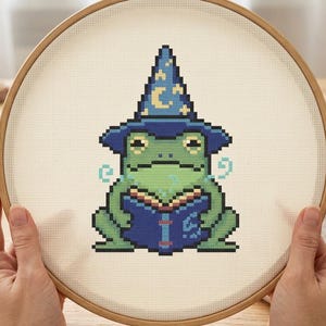 May include: A cross-stitch embroidery project in a round wooden hoop featuring a green frog wearing a dark blue wizard hat with yellow moon and star patterns. The frog is holding a blue book with light blue magical swirls on both sides.