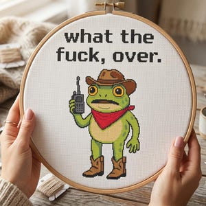 May include: A circular wooden embroidery hoop containing white fabric with a cross-stitch design of a green frog wearing a brown cowboy hat, a red bandana, and brown boots, holding a walkie-talkie. The text reads what the fuck, over.