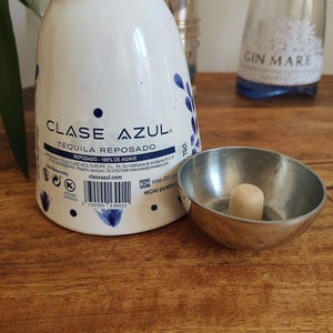 May include: A white Clase Azul Tequila bottle with blue floral designs and the text "TEQUILA REPOSADO". A small metal bowl with a wooden stopper sits next to the bottle. A Gin Mare bottle is in the background.