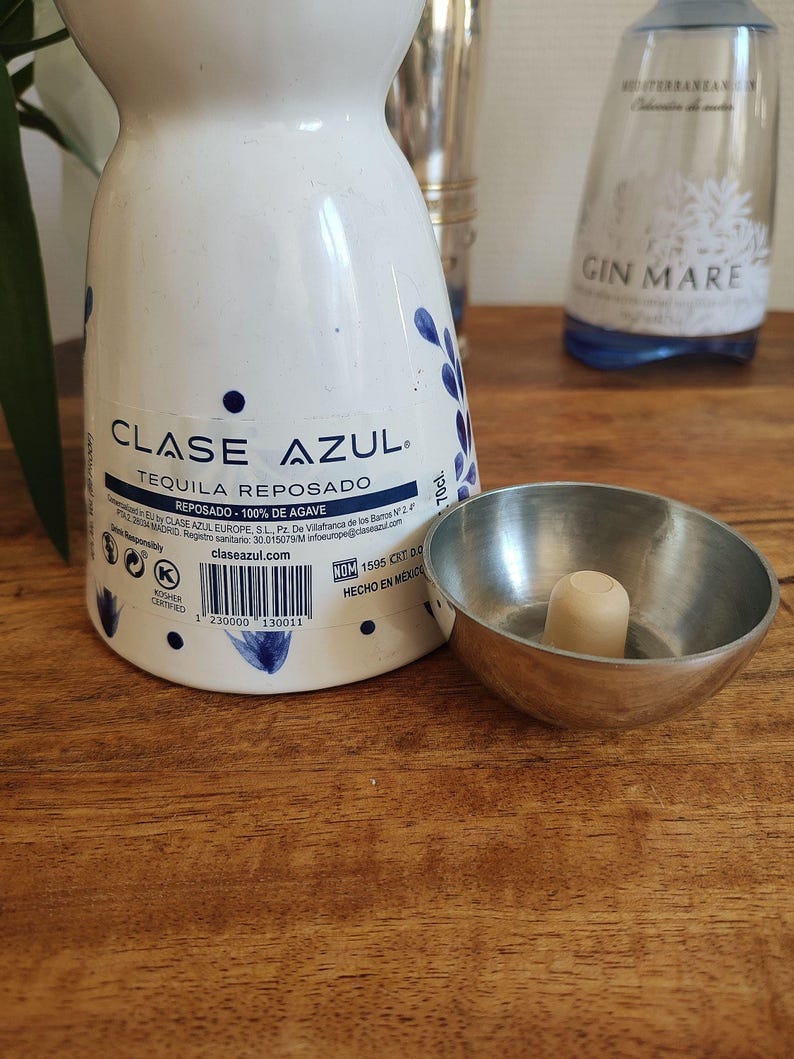 May include: A white Clase Azul Tequila bottle with blue floral designs and the text "TEQUILA REPOSADO". A small metal bowl with a wooden stopper sits next to the bottle. A Gin Mare bottle is in the background.