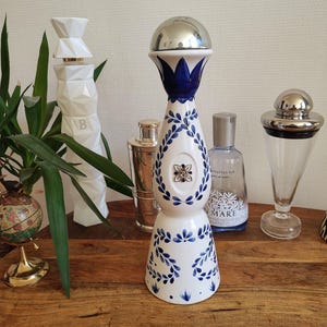 May include: A white and blue ceramic bottle stopper with a silver top. The stopper features a blue floral design and is displayed with other decorative bottles and a globe on a wooden surface.
