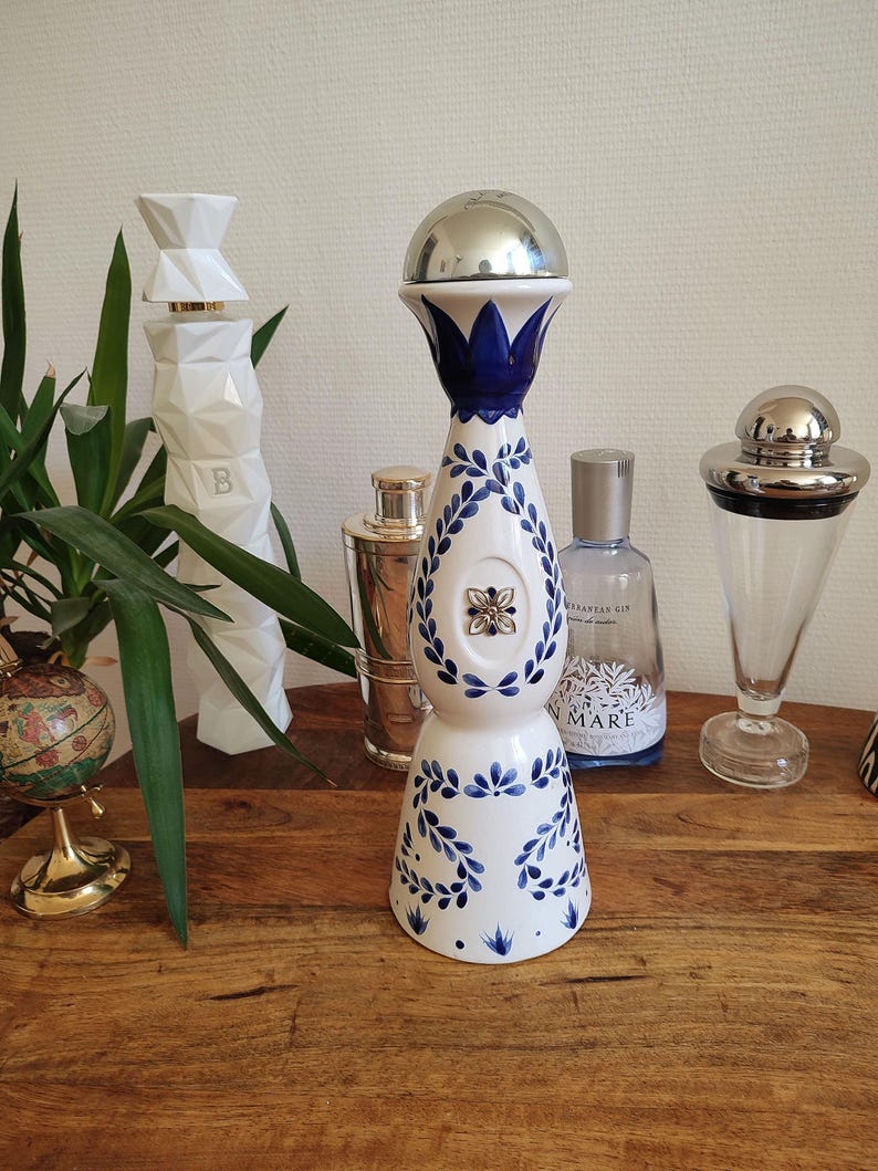 May include: A white and blue ceramic bottle stopper with a silver top. The stopper features a blue floral design and is displayed with other decorative bottles and a globe on a wooden surface.
