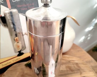 May include: Silver cocktail shaker with a rounded top and a decorative band around the base. The shaker is sitting on a wooden surface.