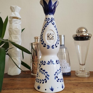 May include: A white ceramic bottle with blue floral designs and a silver top. The bottle has a teardrop shape and is decorated with blue leaves and a silver emblem. Other bottles and a plant are in the background.