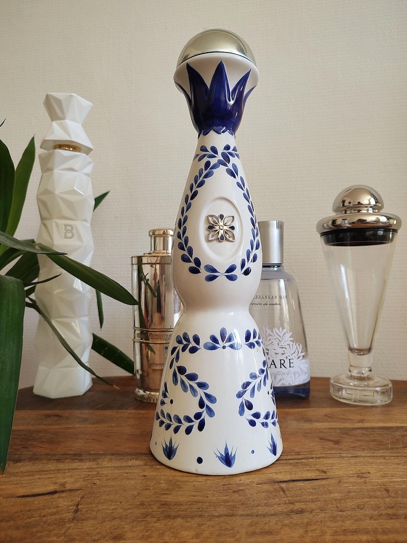 May include: A white ceramic bottle with blue floral designs and a silver top. The bottle has a teardrop shape and is decorated with blue leaves and a silver emblem. Other bottles and a plant are in the background.