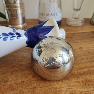 May include: A silver-colored, spherical bottle stopper with the words "CLASE AZUL MEXICO" engraved on its surface. The stopper has a floral design and reflects the surrounding objects. Other bottles and decorative items are in the background.