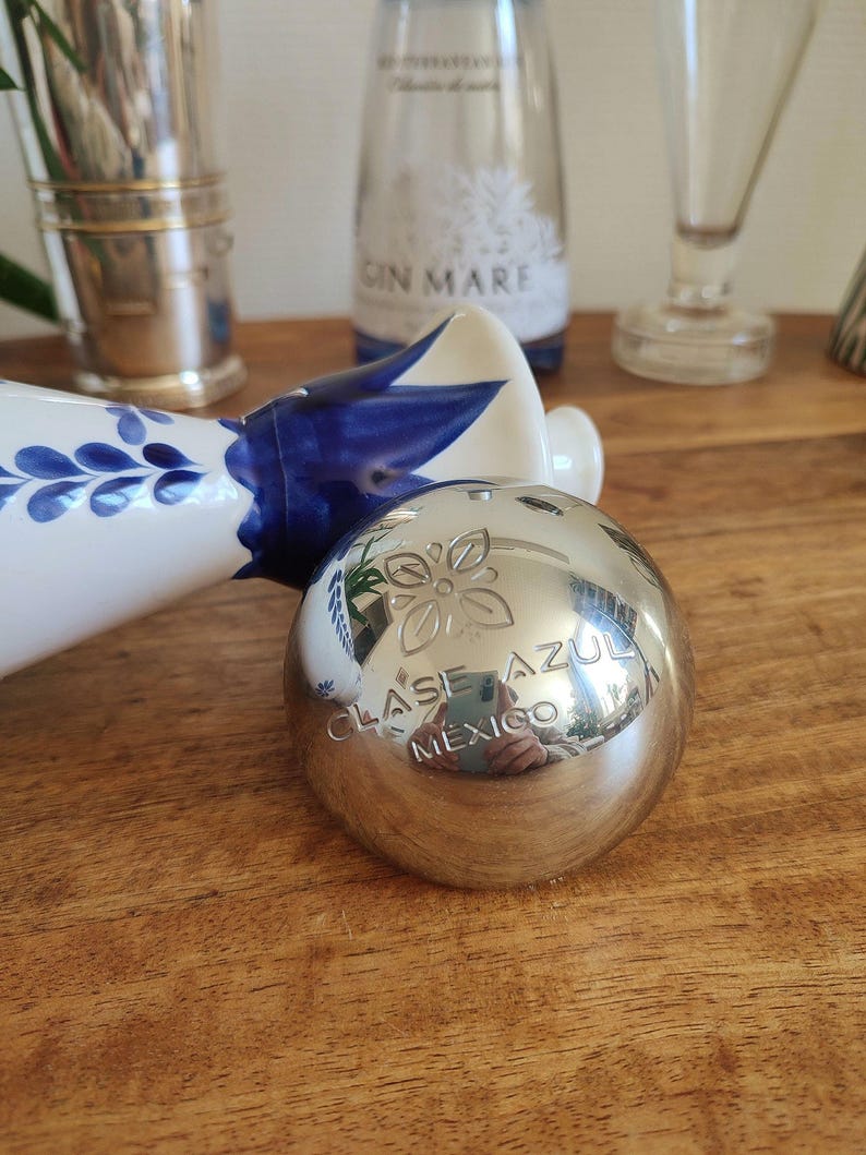 May include: A silver-colored, spherical bottle stopper with the words "CLASE AZUL MEXICO" engraved on its surface. The stopper has a floral design and reflects the surrounding objects. Other bottles and decorative items are in the background.