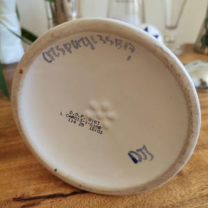 May include: A white ceramic vase base with blue handwritten text and stamped markings. The base has a slightly curved shape and sits on a wooden surface. The text includes numbers and letters.