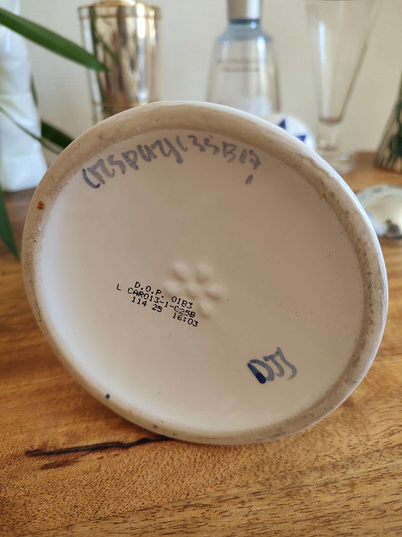 May include: A white ceramic vase base with blue handwritten text and stamped markings. The base has a slightly curved shape and sits on a wooden surface. The text includes numbers and letters.