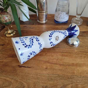 May include: A white ceramic bottle with blue floral patterns and a silver stopper. The bottle is decorated with blue leaves and flowers. Other barware items are in the background.