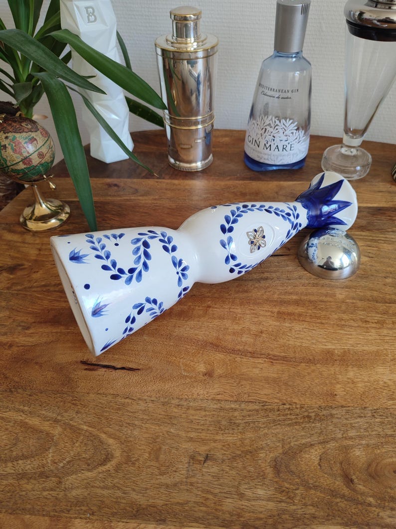 May include: A white ceramic bottle with blue floral patterns and a silver stopper. The bottle is decorated with blue leaves and flowers. Other barware items are in the background.