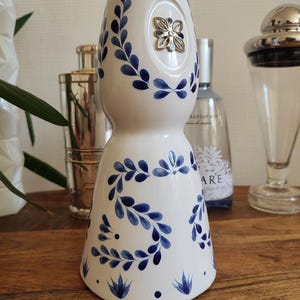 May include: White ceramic bottle with blue floral leaf pattern and a silver flower-shaped embellishment. The bottle has a bulbous shape and a narrow neck. Decorative item for home decor.