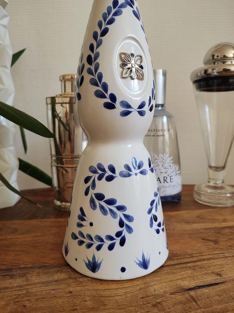 May include: White ceramic bottle with blue floral leaf pattern and a silver flower-shaped embellishment. The bottle has a bulbous shape and a narrow neck. Decorative item for home decor.