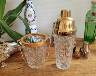 May include: A gold-rimmed, cut-glass ice bucket with a handle sits next to a gold-topped cocktail shaker. A decorative ceramic bottle with blue floral designs and a green glass bottle are also visible. A collection of barware.