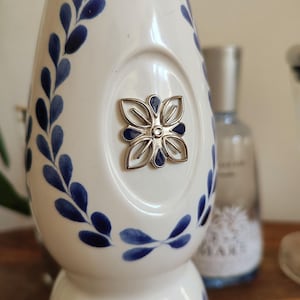 May include: White ceramic bottle with blue floral leaf pattern and a silver flower emblem. The bottle has a rounded shape and a narrow neck. The design is reminiscent of Delftware pottery.