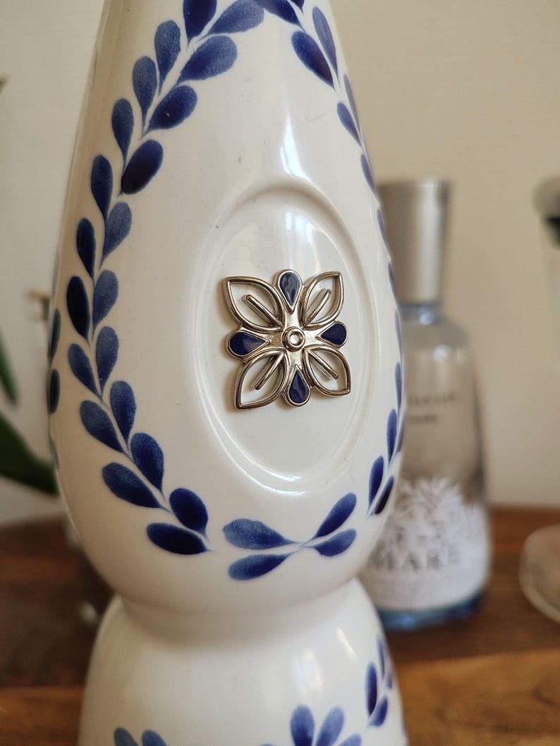 May include: White ceramic bottle with blue floral leaf pattern and a silver flower emblem. The bottle has a rounded shape and a narrow neck. The design is reminiscent of Delftware pottery.