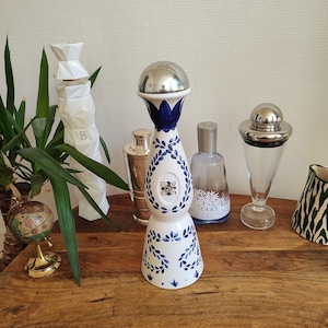 May include: A selection of decorative bottles and barware on a wooden surface. The focal point is a white and blue ceramic bottle with a silver top. Other items include a cocktail shaker, a glass decanter, and a globe.