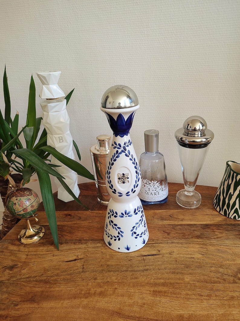 May include: A collection of decorative bottles and barware on a wooden surface. The centerpiece is a white and blue ceramic bottle with a silver top. Other items include a cocktail shaker, a glass decanter, and a globe.