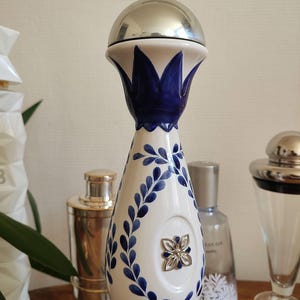 May include: A decorative bottle with a silver top and a white ceramic body. The bottle is adorned with blue floral patterns and a silver flower emblem. The design is reminiscent of traditional Mexican pottery.