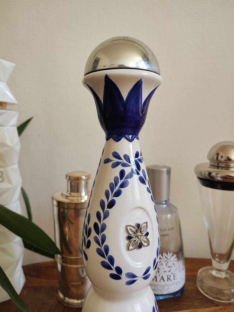May include: A decorative bottle with a silver top and a white ceramic body. The bottle is adorned with blue floral patterns and a silver flower emblem. The design is reminiscent of traditional Mexican pottery.