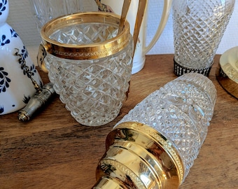 May include: A collection of barware including a crystal ice bucket with a gold rim and handle, a crystal cocktail shaker with a gold cap, and two additional glass shakers with gold accents, one featuring a decorative illustration of a person.