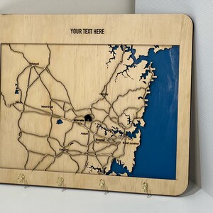 Può includere: Portachiavi in legno con una mappa di Sydney, Australia. La mappa è tagliata al laser e presenta le principali strade e punti di interesse della città. Il portachiavi ha uno sfondo blu e una cornice in legno. Il testo "YOUR TEXT HERE" è stampato nella parte superiore della cornice.