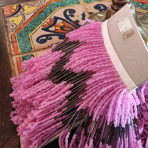 May include: Close-up of a craft project featuring strands of pink beads attached to a cardboard base. The beads are a vibrant pink color, and the project is resting on a decorative tile surface.