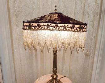 May include: A gold table lamp with a burgundy and gold patterned lampshade with a beaded fringe. The lamp is on a small table with a floral pattern.