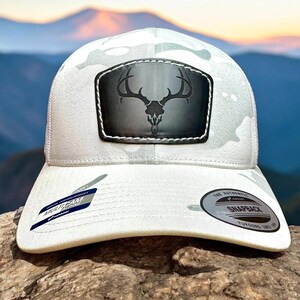 May include: White camouflage trucker hat with a black leather patch featuring a skull with antlers. The hat has a black snap closure and a white mesh back.