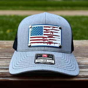 May include: A gray and black trucker hat with an American flag design featuring a row of guns. The flag is on a metal patch with silver rivets. The hat has a black mesh back and a Richardson 112 Original Trucker label.