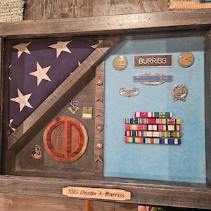 May include: A shadow box display with a folded American flag, military medals, and ribbons. The display includes a wooden emblem and a nameplate that reads "SSG Dustin A. Burriss". The frame is made of dark wood.