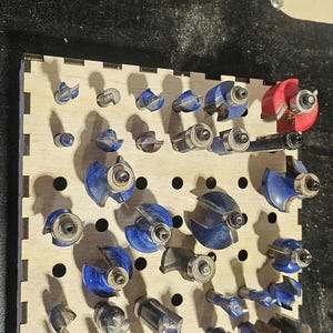 Puede incluir: Un organizador de madera con una rejilla de agujeros que contiene varias brocas de enrutador. Las brocas son una mezcla de plata, azul y rojo, mostrando diferentes perfiles de corte. El organizador es de madera clara.