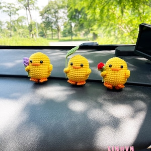 May include: Three yellow crochet chick figurines with orange feet and black eyes. Each chick holds a small flower or leaf. The figurines are displayed on a dark surface, with the text "XINHVN" visible.